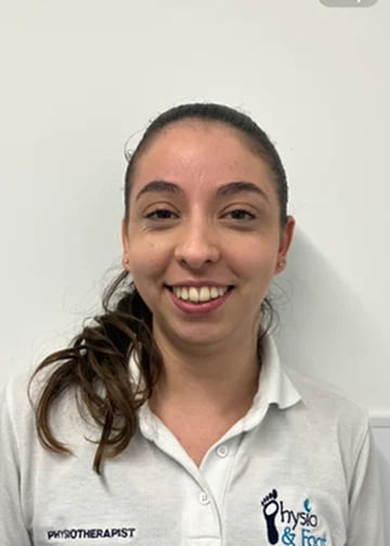 Professional headshot of a smiling woman with dark hair in a ponytail, wearing a white physiotherapist uniform